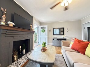 A living room with a fireplace, a television, a ceiling fan, a door with a wreath, and a sofa with colorful cushions.