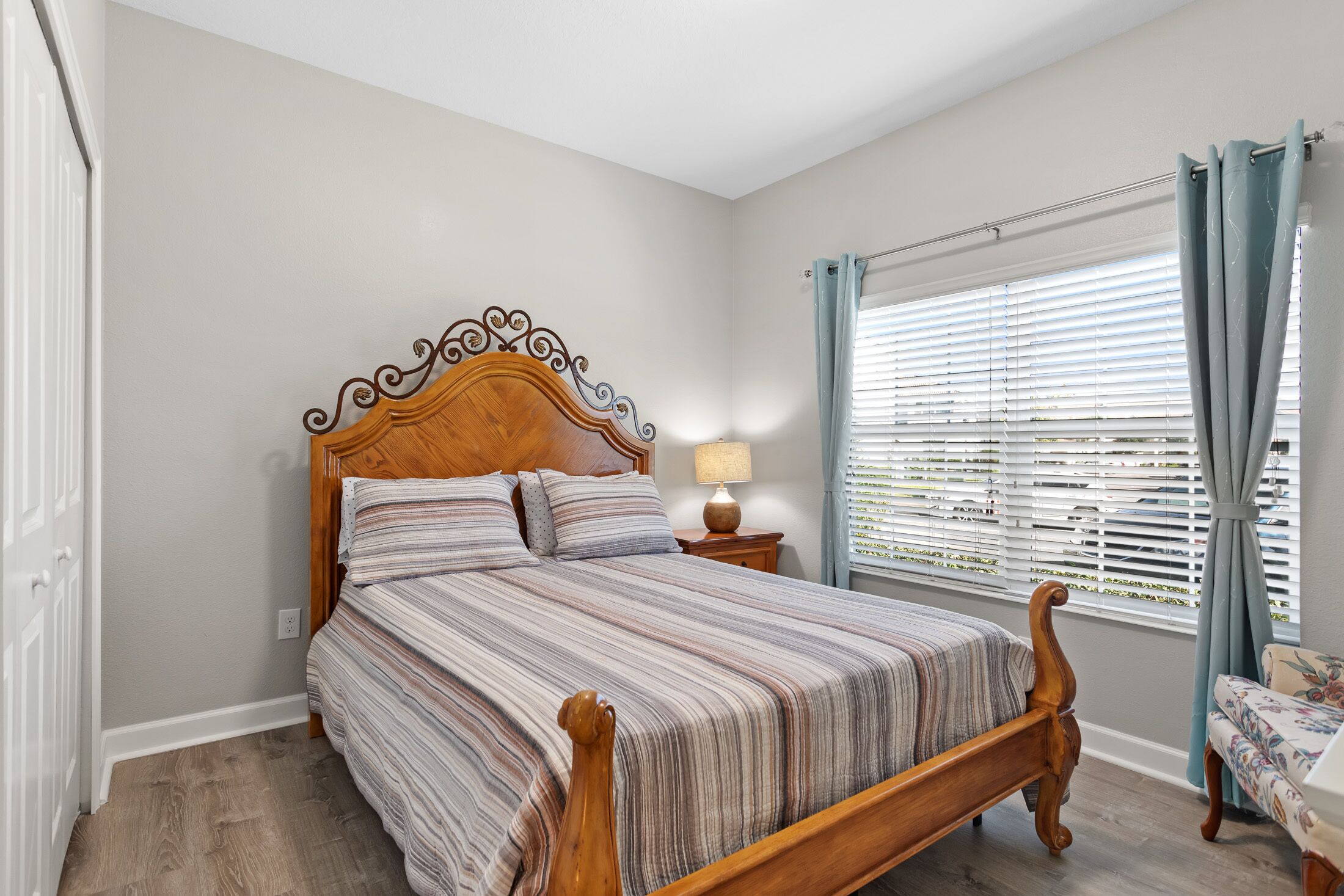 A bedroom with a wooden bed, a nightstand, a lamp, and a window with blinds.