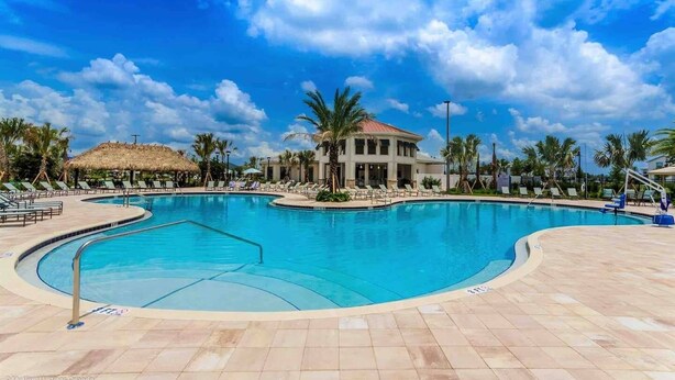 A large outdoor swimming pool with lounge chairs and palm trees, surrounded by a building with a thatched roof.