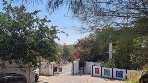 A paved courtyard with a car parked on the left, a person standing near a wall, and a gate with colorful signs. - Nature Retreat Cottages Jaisamand Udaipur  (Udaipur)