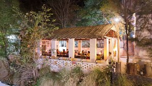 A rustic outdoor restaurant with a thatched roof, surrounded by trees and vegetation. - Nature Retreat Cottages Jaisamand Udaipur  (Udaipur)