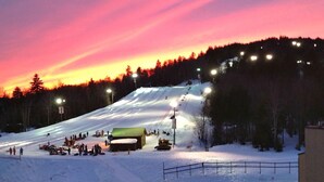 Ein mit Schnee bedeckter Skipisten im Dämmerlicht mit künstlicher Beleuchtung und einem kleinen Gebäude unten.