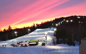 A snow-covered ski slope at dusk with artificial lighting and a small building at the bottom.