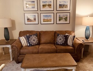 A brown leather sofa with decorative pillows, a matching ottoman, and framed landscape paintings on the wall.