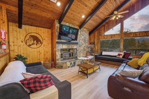 A cozy cabin interior with a stone fireplace, wooden walls, and large windows offering a view of the outdoors.