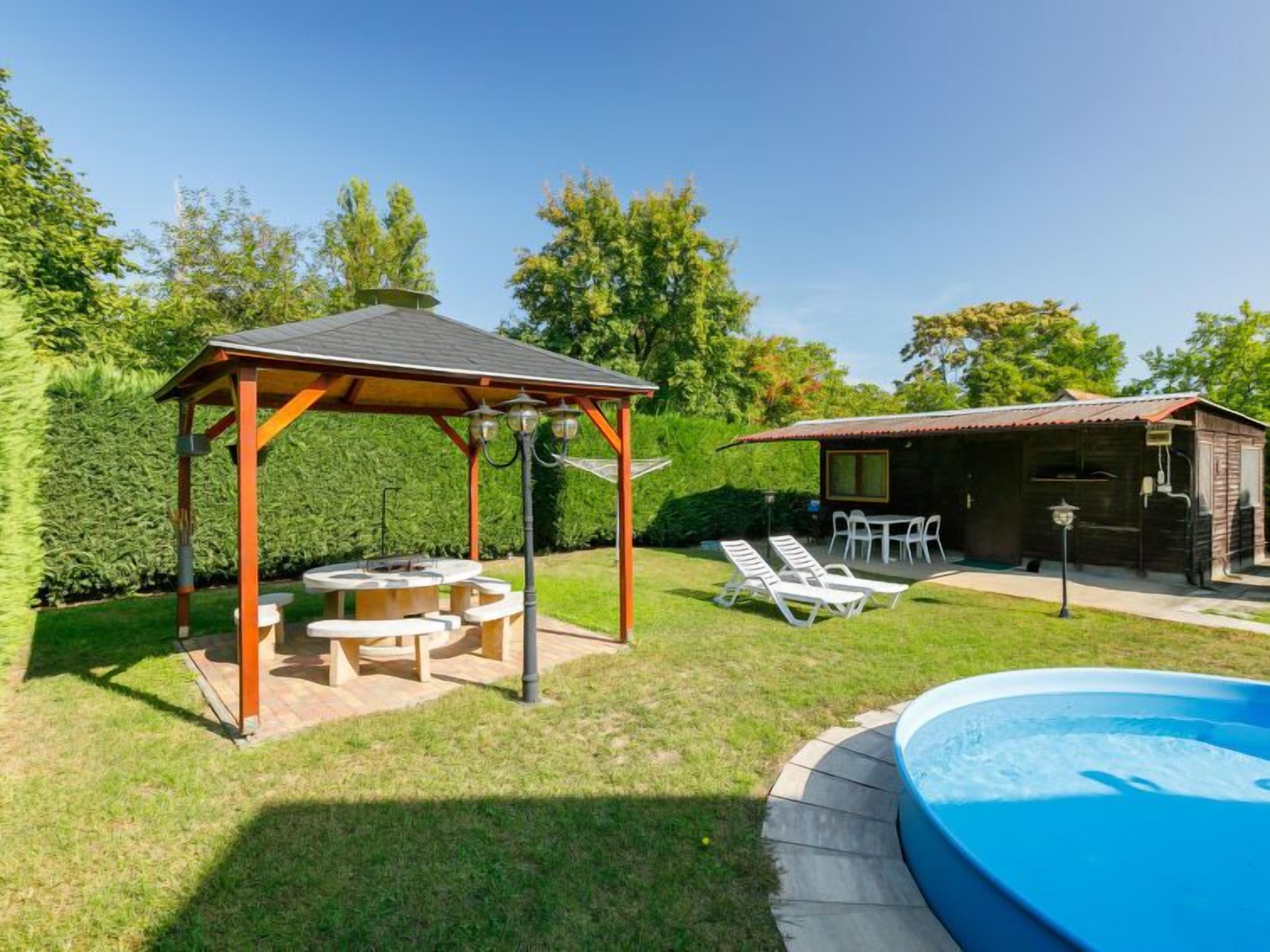 A backyard with a wooden gazebo, outdoor seating, a barbecue area, a pool, and a small cabin-like structure.