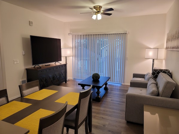 A modern living room with a dining area, a flat-screen TV, a ceiling fan, and a large window with blinds. - #203 New 2BR Condo near Evergreen State Fairgrounds (Monroe)
