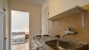 A compact kitchen with a sink, stove, and a small countertop. A sliding door leads to a bedroom with a bed and a nightstand. - Vacation home Edina by Interhome (Balatonszarszo)