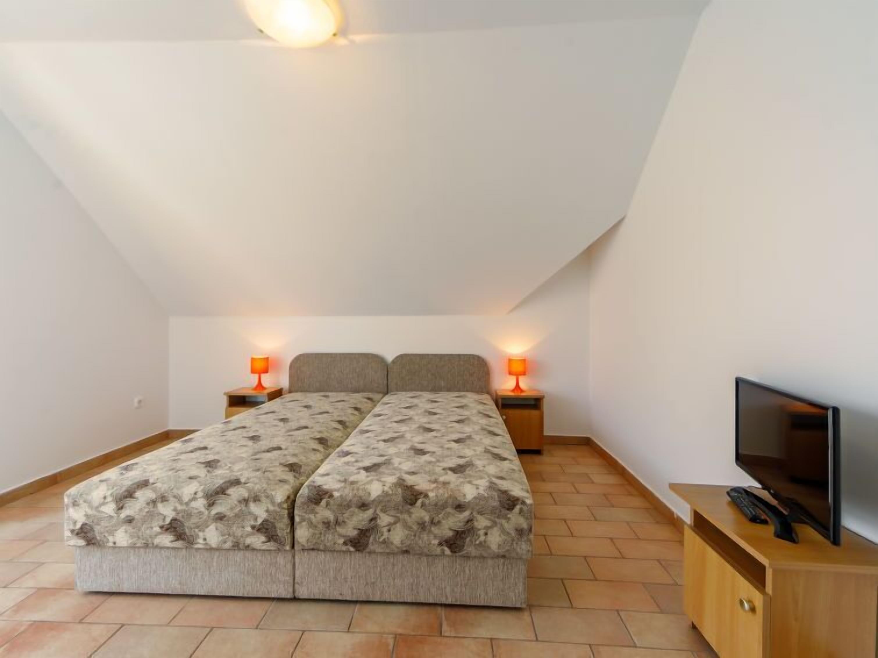 A bedroom with a bed, two bedside lamps, a TV on a wooden stand, and a sloped ceiling.