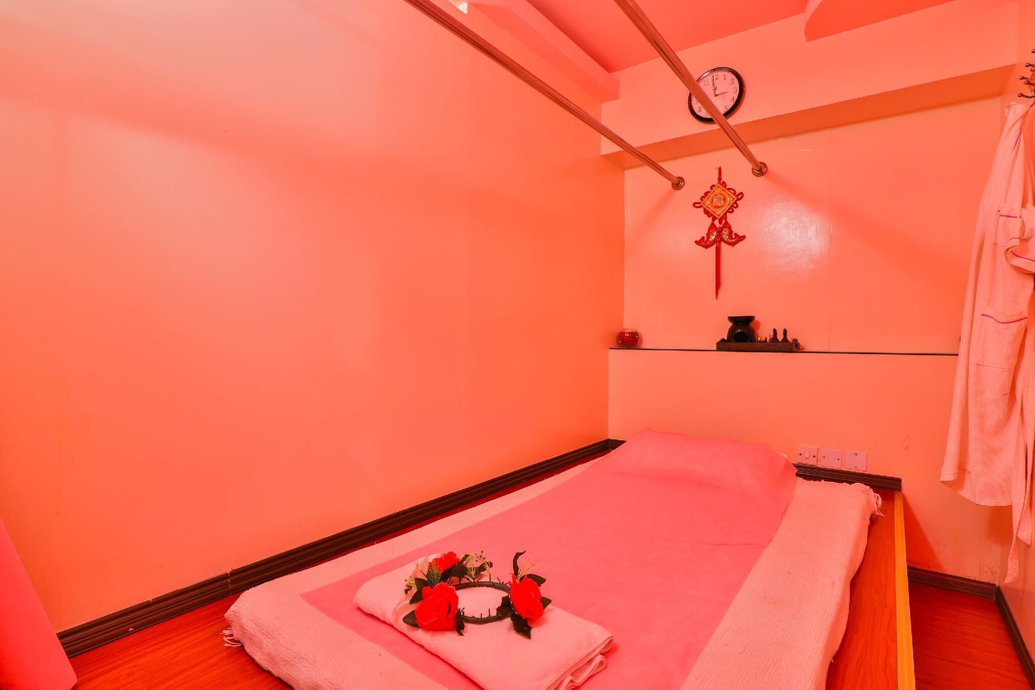 A single bed with a pink towel and a floral headband, a clock on the wall, and a robe hanging on the right.