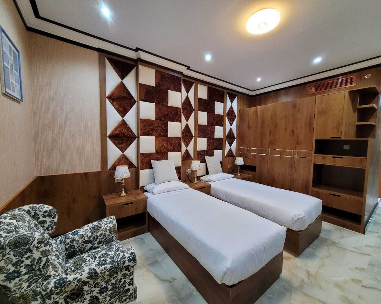 Habitación de hotel con dos camas, cabeceros de madera, un sillón estampado y un armario empotrado.