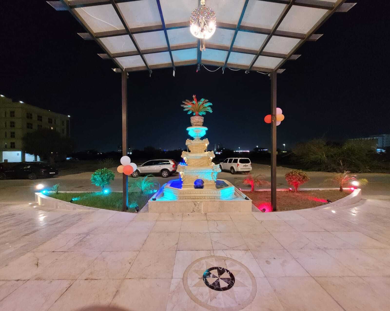 A well-lit outdoor area with a decorative fountain, a covered seating area, and a paved walkway.