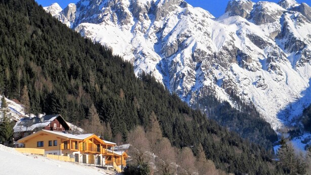 Un chalet de montagne à l’extérieur en bois, entouré de sommets enneigés et d’une forêt dense.