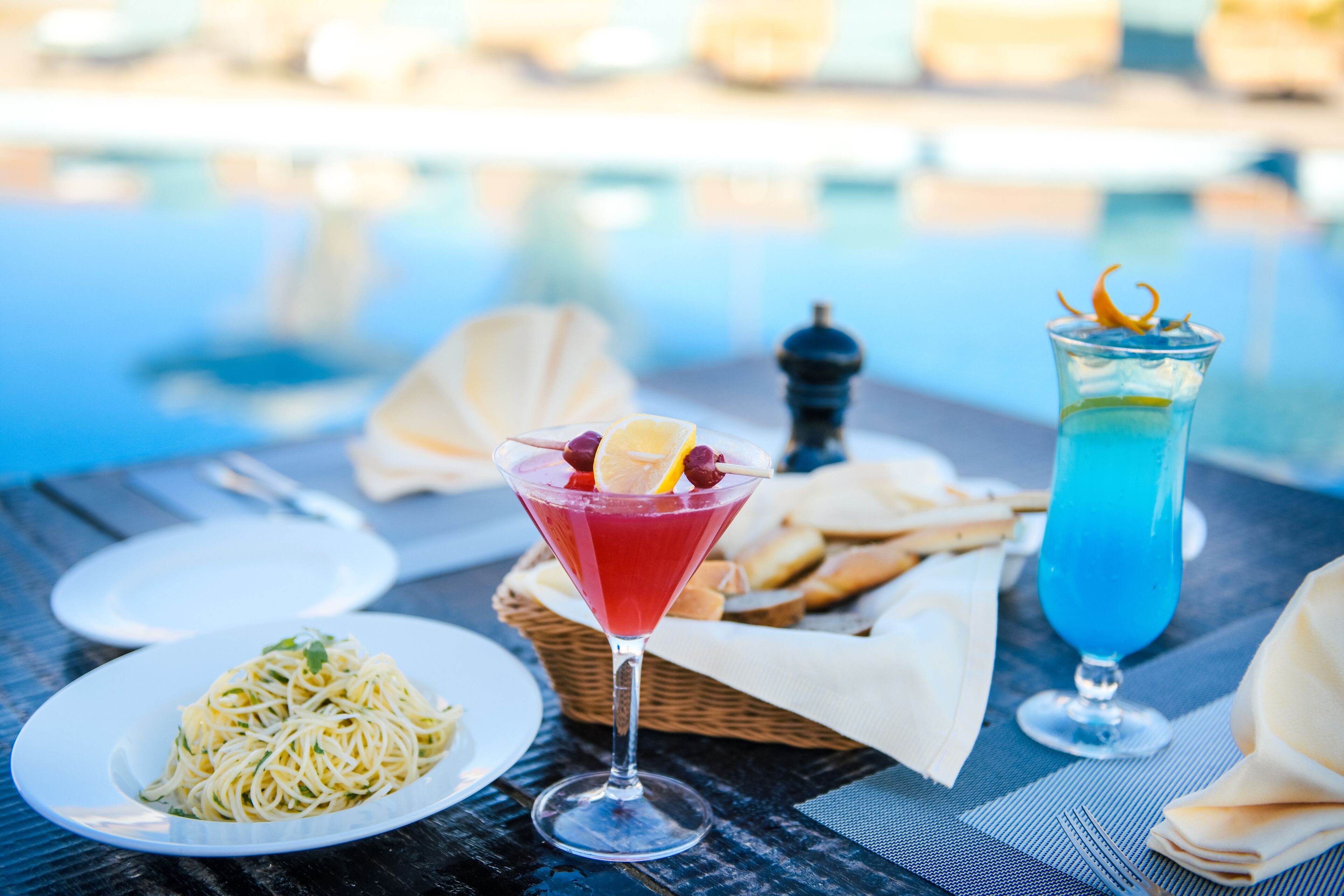 A dining table with a plate of spaghetti, a martini glass with a red drink, and a tall glass with a blue drink.