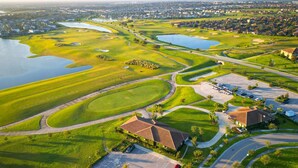 En vy från ovan av en golfbana med flera vattenhinder, ett klubbhus och bostadsområden i bakgrunden.