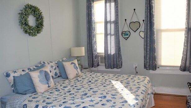 A bedroom with a bed, a nightstand, a lamp, a window with curtains, and wall decorations including a wreath and heart-shaped mirrors. - OCEAN BLOCK Downtown Rehoboth Beach 2BR Condo Sleeps 6 (Rehoboth Beach)