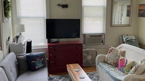 A living room with a grey sofa, a red dresser, a wooden coffee table, a white wicker sofa, and a TV on a stand. - OCEAN BLOCK Downtown Rehoboth Beach 2BR Condo Sleeps 6 (Rehoboth Beach)