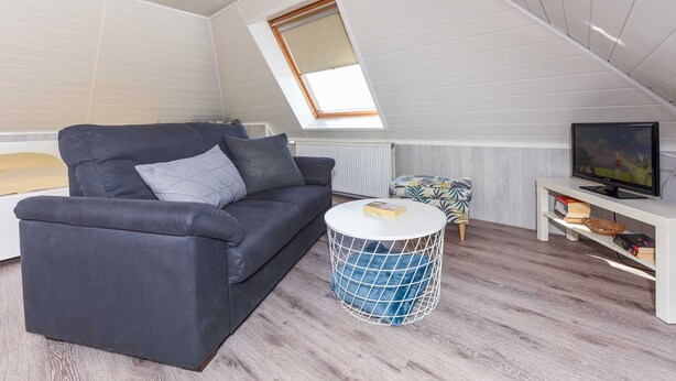Quarto moderno estilo loft com teto inclinado, sofá cinza escuro, mesa de centro redonda e uma pequena mesa com televisão, além de um radiador.