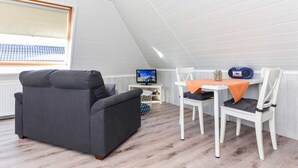 Um quarto aconchegante com um sofá cinza escuro, uma mesa de jantar branca com duas cadeiras, uma televisão sobre um móvel pequeno e uma janela com persianas.