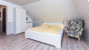 Quarto com teto inclinado, cama com roupa de cama amarela, guarda-roupa branco e poltrona estampada.