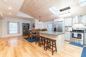 A modern kitchen with wooden flooring, a central island with stools, stainless steel appliances, and a skylight.