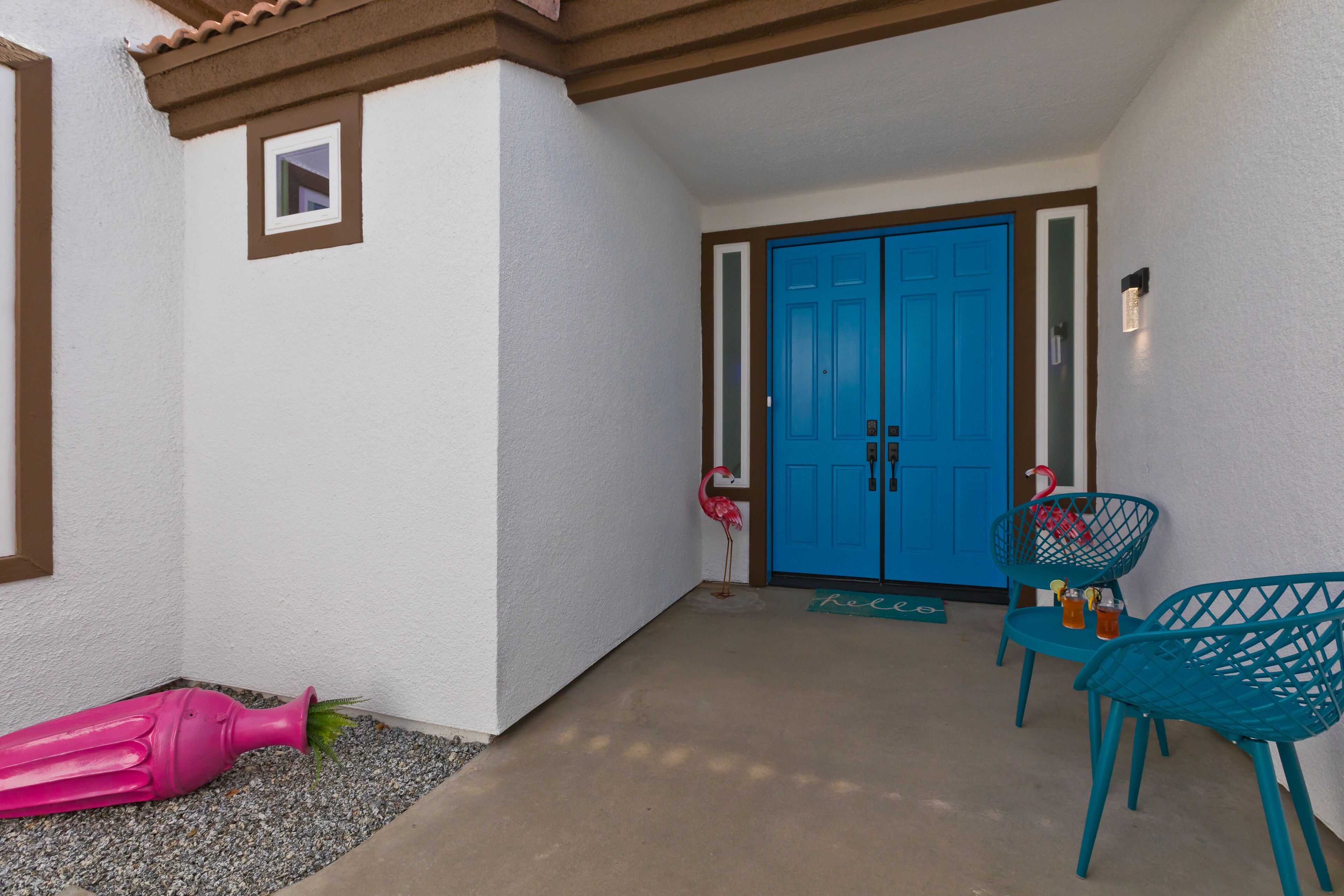 A covered patio with a blue door, two turquoise chairs, and a pink flamingo decoration.