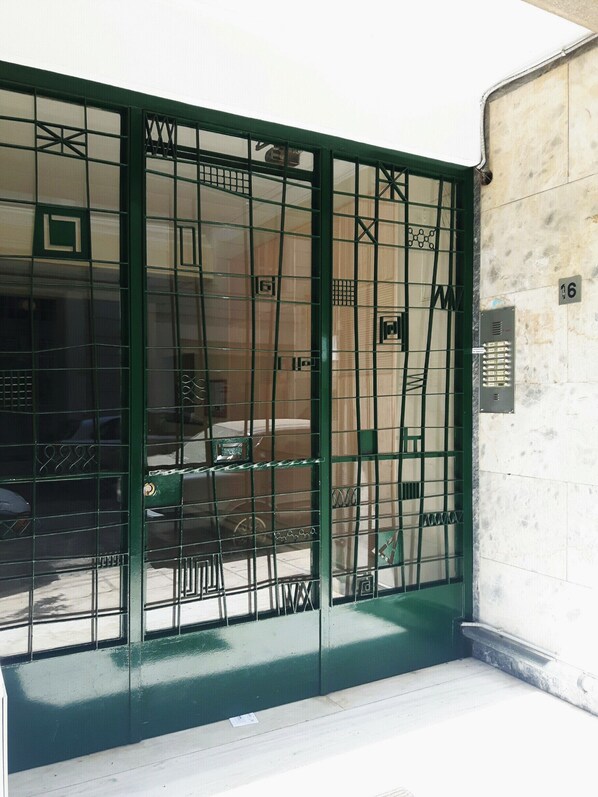 Une porte métallique verte ornée d’un motif en treillis, un mur blanc avec une plaque numérotée et un petit espace extérieur comprenant une voiture et une table.