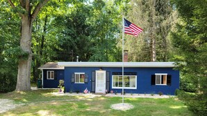 Uma casa térrea com fachada azul, um mastro com a bandeira americana e um pequeno jardim frontal com grama e uma árvore.