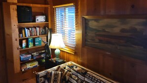 Um quarto aconchegante com paredes de madeira, uma estante cheia de livros, uma escrivaninha com luminária e uma janela com persianas.