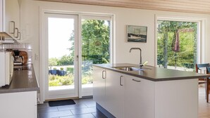 A modern kitchen with a large island, a built-in sink, and a coffee maker. There are two large windows providing views of greenery and a pathway. - Stunning home in Allinge (Allinge)