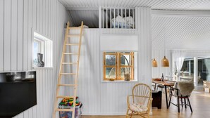 Ett rum med en säng på ett loft, en trästege, en fåtölj i rotting och en matplats med ett bord och stolar.
