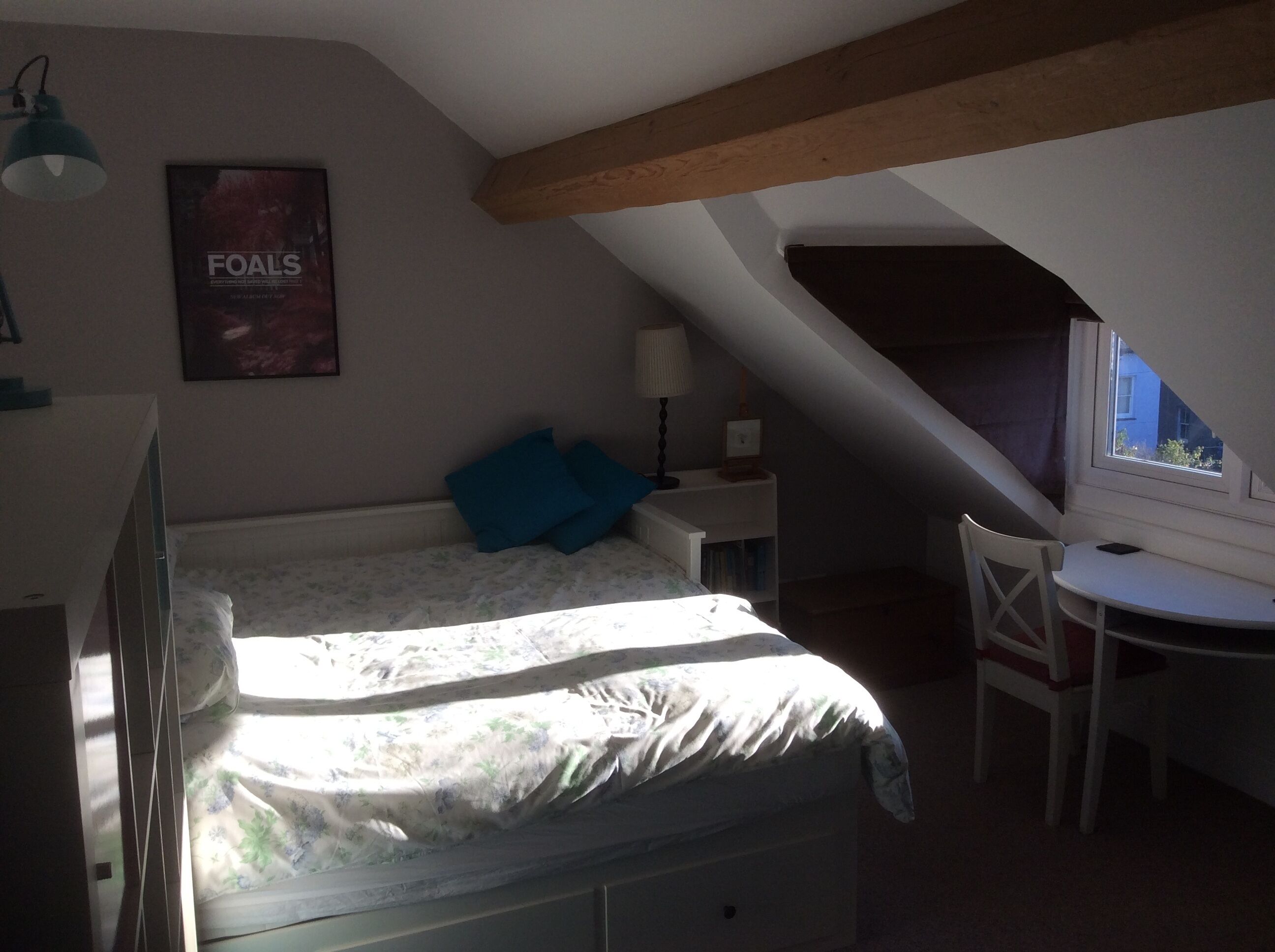 Un dormitorio con cama, escritorio, silla, lámpara, un cartel en la pared y ventana.