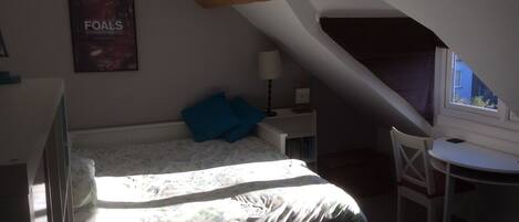 Un dormitorio con cama, escritorio, silla, lámpara, un cartel en la pared y ventana.