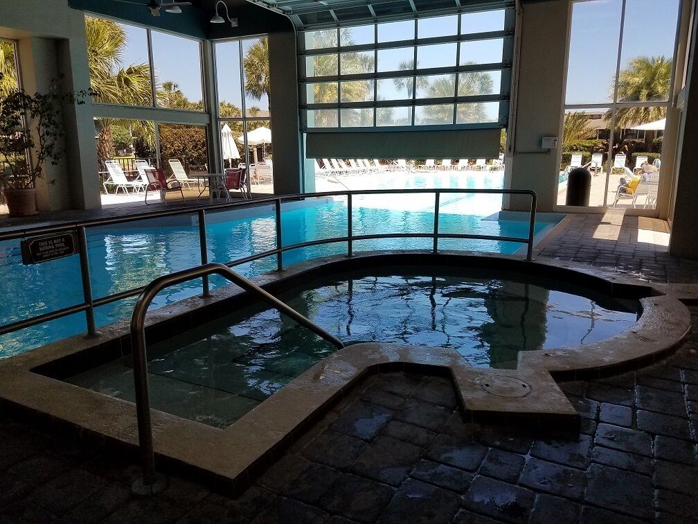 Piscina interior com paredes de vidro transparente, chão ladrilhado e vista para a área da piscina exterior.