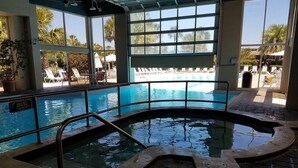 Una piscina interior con paredes de vidrio transparente, suelo de baldosas y vista al área de la piscina exterior.