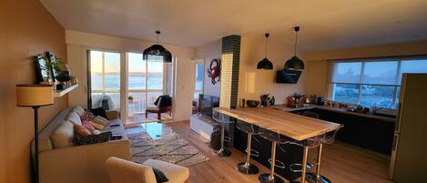A modern open-plan living and dining area with a large window, a sofa, a coffee table, and a bar area with stools.