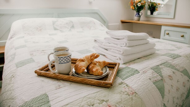 En snyggt bäddad säng med ett täcke, ett bricka med croissanter och kaffekoppar och en hög med handdukar.