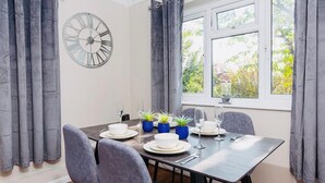 A dining area with a table set for four, including plates, bowls, glasses, and cutlery. There are four chairs, a clock on the wall, and a window with curtains. - Beautiful 1-bed Apartment in York Near Gym (York)