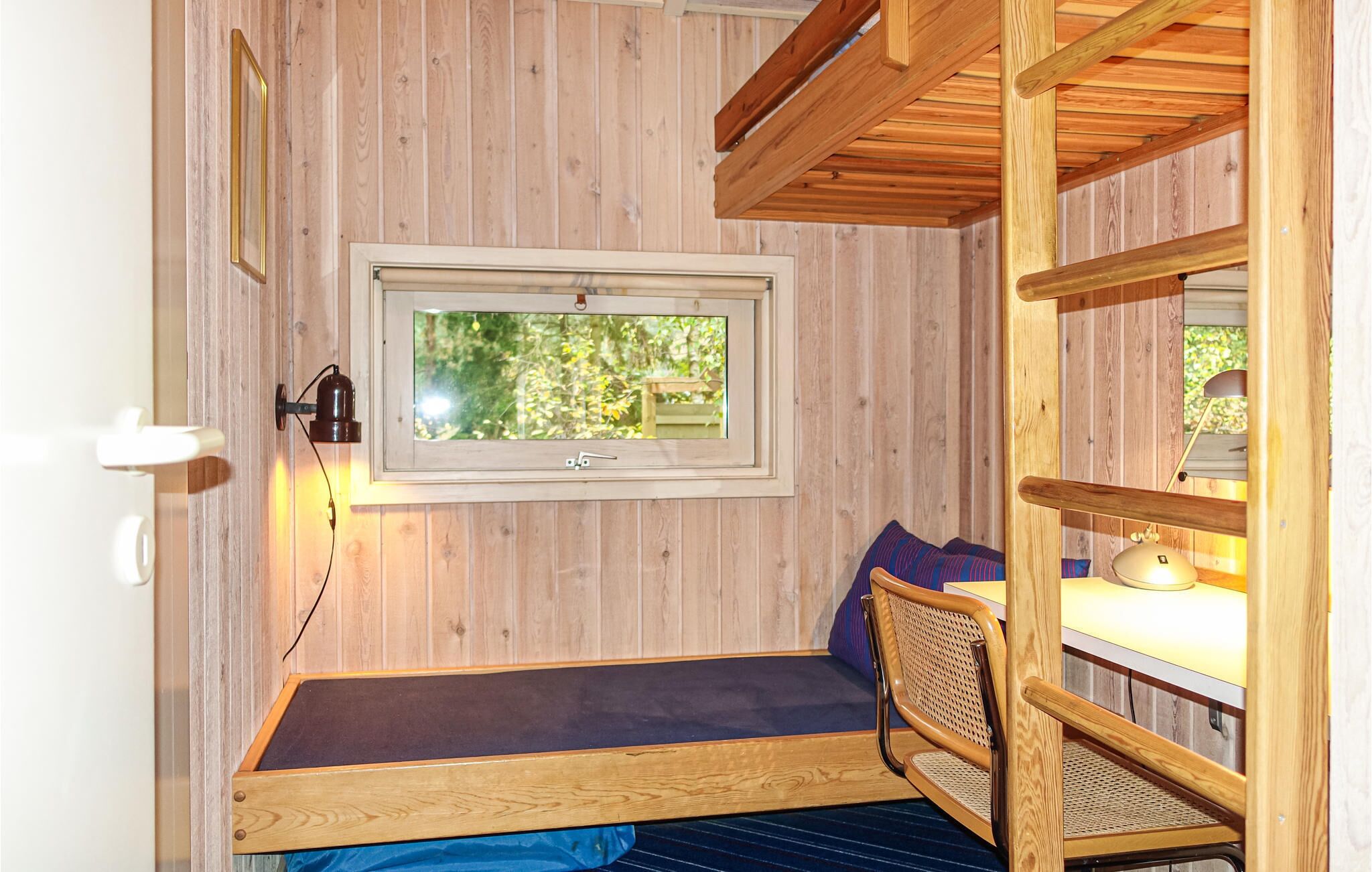 Una stanza modesta con travi in legno al soffitto, una finestra che si affaccia sul verde, un letto con materasso scuro, una sedia e una lampada.