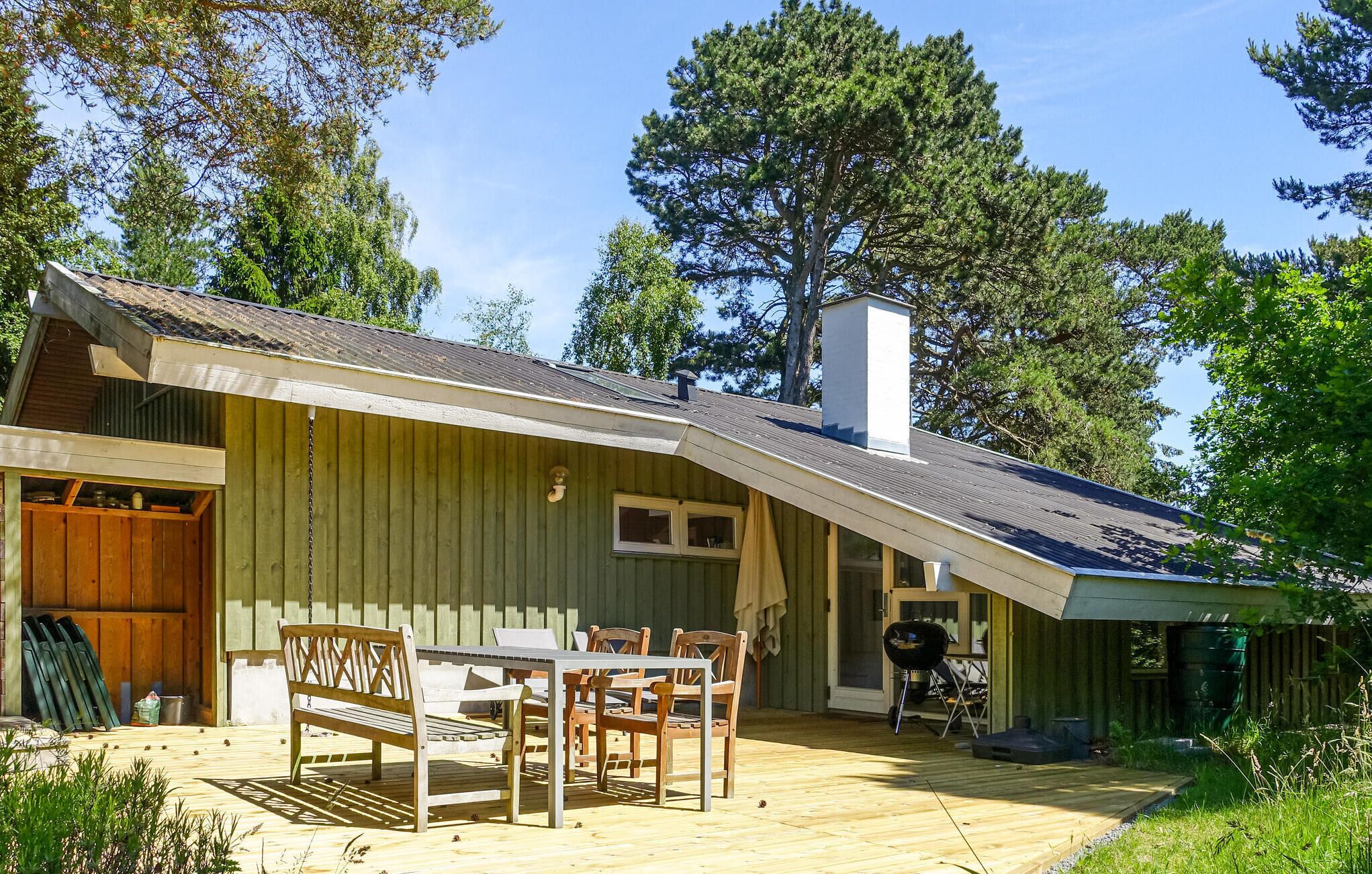 A wooden cabin with a deck, a dining area with chairs and a table, and a chimney.