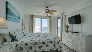 A bedroom with a bed, a ceiling fan, a TV, and a view of the sea.