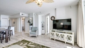 A spacious living room with a large flat-screen TV, a dining area with a wooden table and chairs, and a cozy seating area with a wicker chair.