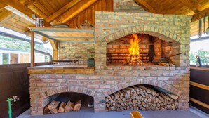 Forno de pedra a lenha com fogo aceso, uma pilha de lenha embaixo e uma prateleira de metal acima com utensílios de cozinha.