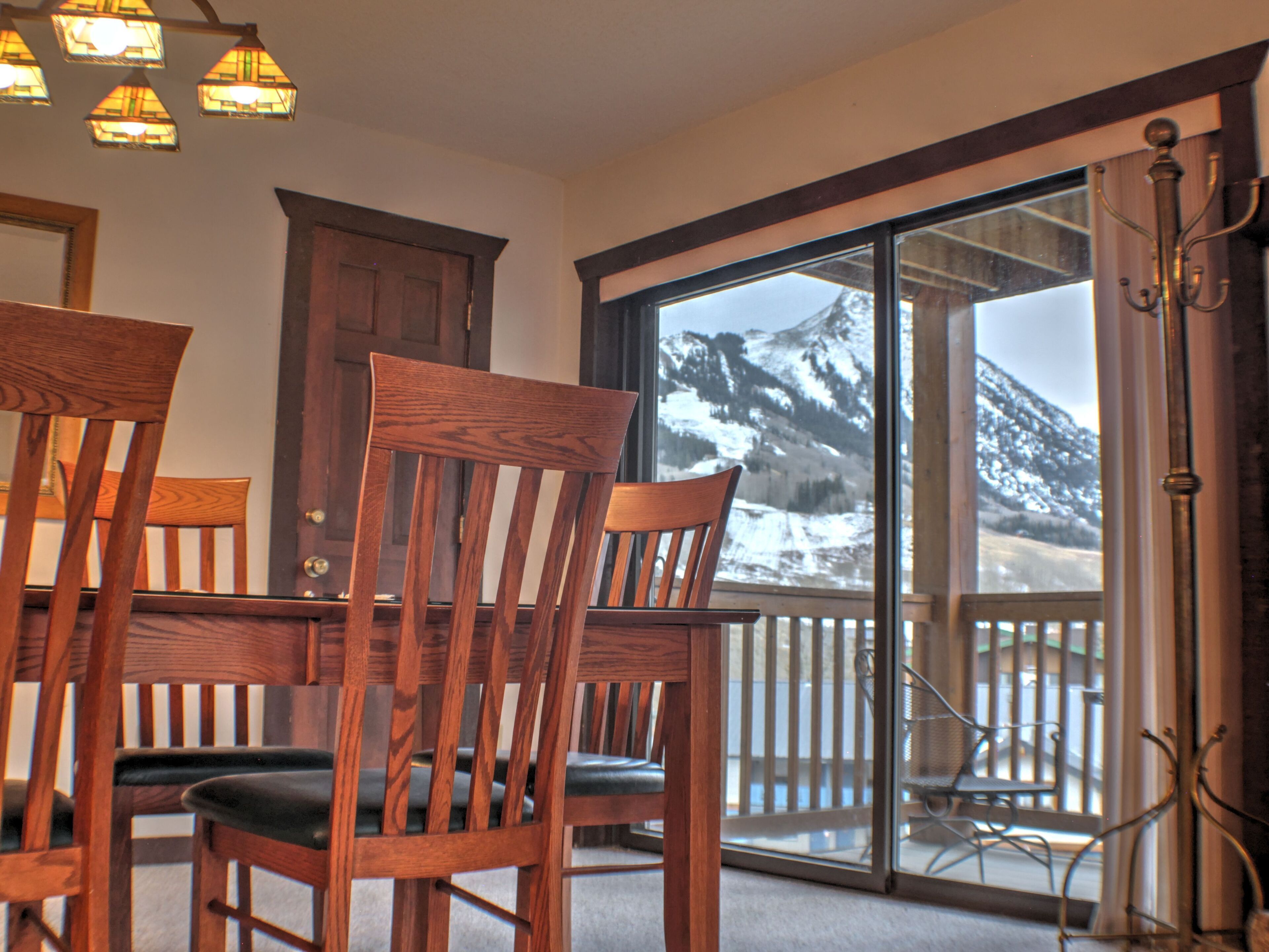 A dining area with wooden chairs and a table, a wooden door, and a large sliding glass door leading to a balcony with a mountain view.