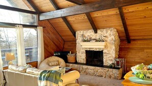 A cozy living room with a stone fireplace, wooden ceiling beams, a comfortable sofa, and a view through a large window. - Bel-Hygge 🐋Chalet, cozy comfort the AK way (Anchorage)