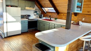 A kitchen with wooden beams, a refrigerator, stove, sink, and a trash bin. - Bel-Hygge 🐋Chalet, cozy comfort the AK way (Anchorage)