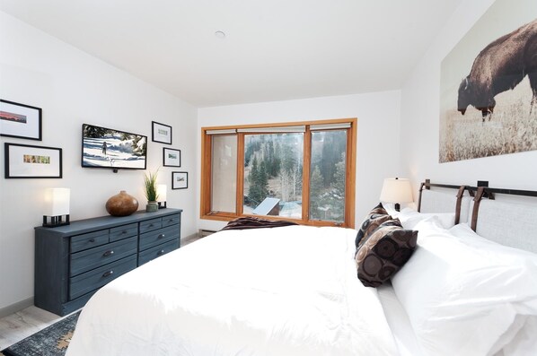 A bedroom with a large bed, a dresser, a window with a view of trees, and framed pictures on the wall. - Modern Village Center Ski In/Out Condo with Slope Views (Durango)