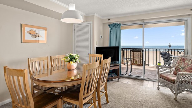 Een eethoek met een ronde tafel en stoelen, een televisie, en een balkon met uitzicht op het strand.