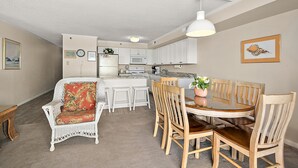 Een kleine keuken met witte kasten, een magnetron en een koelkast. Er is een eethoek met een ronde tafel en houten stoelen.