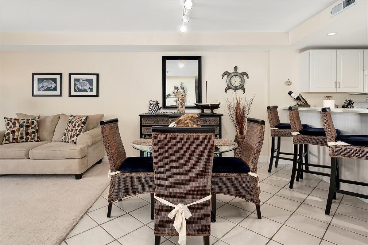 A modern living room with a sofa, dining table, and chairs. There is a kitchen area with cabinets and a clock on the wall.